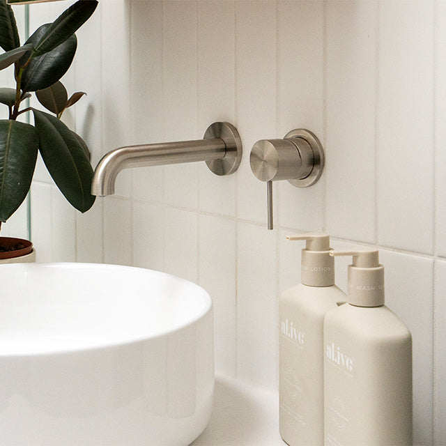 Nero Mecca wall-mounted basin/bath spout in brushed nickel, shown over a white basin with matching wall outlet in a bathroom.