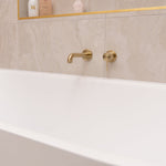 Nero Mecca wall-mounted basin/bath spout in brushed gold above a white bath, with round gold mixer control on tiled wall
