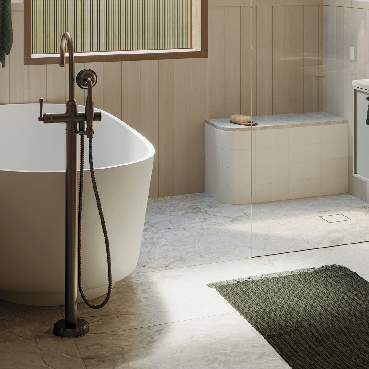 Phoenix Cromford floor-mounted bath mixer with hand shower in brushed bronze beside a freestanding bath in a modern bathroom