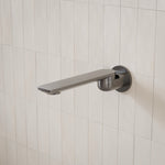 Caroma Urbane II round wall-mounted bath spout in gunmetal finish, shown against white tiled wall at side angle