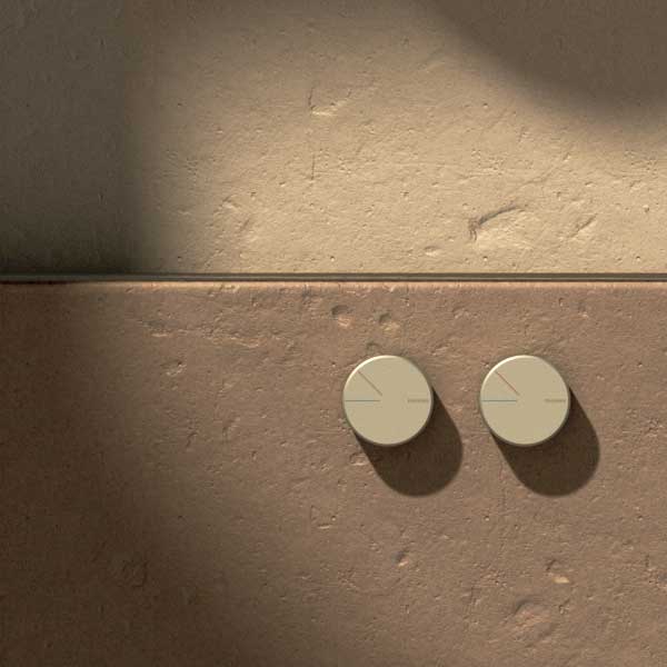 Two brushed nickel Phoenix Axia wall mixer plates shown side by side on a tiled bathroom wall, round and minimalist