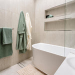 Bao Elegant white back-to-corner bath in a tiled bathroom, shown from the front-right corner with glass screen and towels