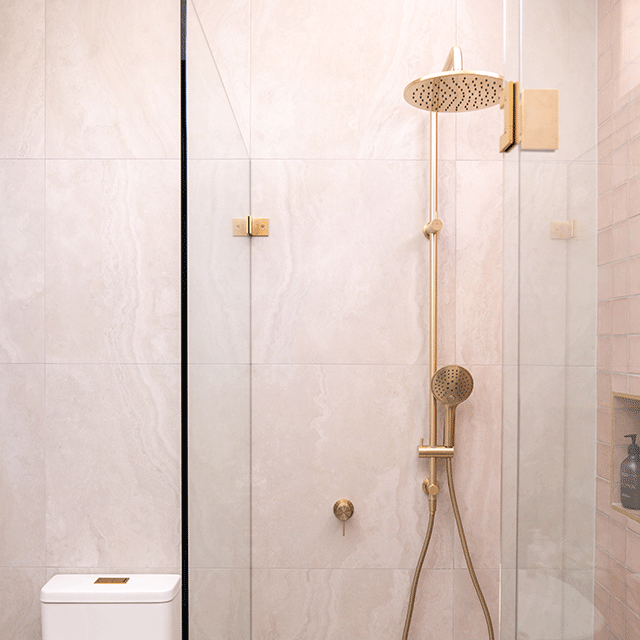 Brushed gold Nero Mecca wall shower mixer on marble-look bathroom tiles with matching shower set and toilet nearby