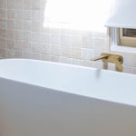 Caroma Contura II wall basin/bath mixer in brushed brass, shown mounted above a white bath against tiled wall.