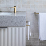 Caroma Contura II brushed brass single towel rail holding a white towel in a tiled bathroom, side view