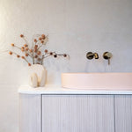 Caroma Liano II wall-mounted bath/shower mixer in brushed brass, shown above a pink basin in a modern bathroom