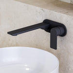 Caroma Urbane II wall-mounted basin mixer in matte black, shown over a white basin against stone tiles