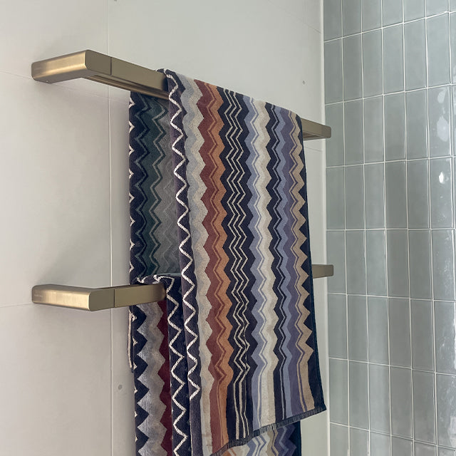 Caroma Urbane II double towel rail in brushed brass, mounted on a bathroom wall with two striped towels