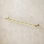 Caroma Urbane II double towel rail in brushed brass, wall-mounted on a light stone bathroom tile wall