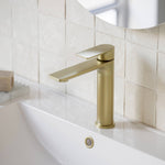 Caroma Urbane II mid tower basin mixer in brushed brass, shown mounted on a bathroom vanity beside a sink.