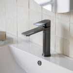 Caroma Urbane II mid tower basin mixer in gunmetal, hob mounted on a white basin beside tiled bathroom wall