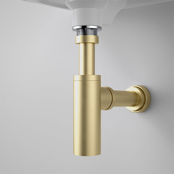 Caroma bottle trap in brushed brass, shown under a wall-hung basin with exposed waste connection