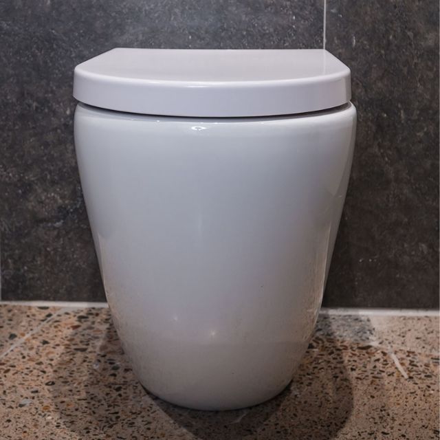 Fienza Aluca back-to-wall toilet suite in gloss white with slim closed seat, front view against dark tiled wall.