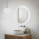 Ingrain round frameless backlit LED mirror above bathroom vanity, shown lit in a modern white tile setting