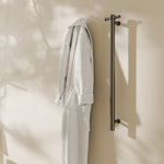 Nero Mecca single vertical heated towel rail in gun metal, wall mounted with white robe in a bathroom setting