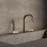 Nero Serenity three-piece basin set in brushed bronze with travertine handles, shown in a modern concrete bathroom setting