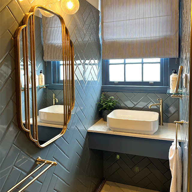 Phoenix Cromford double towel rail in brushed gold, mounted on a blue bathroom wall beside a vanity and mirror