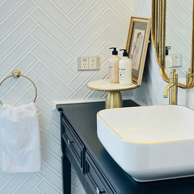 Phoenix Cromford brushed gold hand towel holder with towel mounted on a bathroom wall beside a basin sink