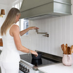 Phoenix Cromford wall mounted pot filler in brushed nickel above a cooktop, shown in a kitchen with a woman filling a pot