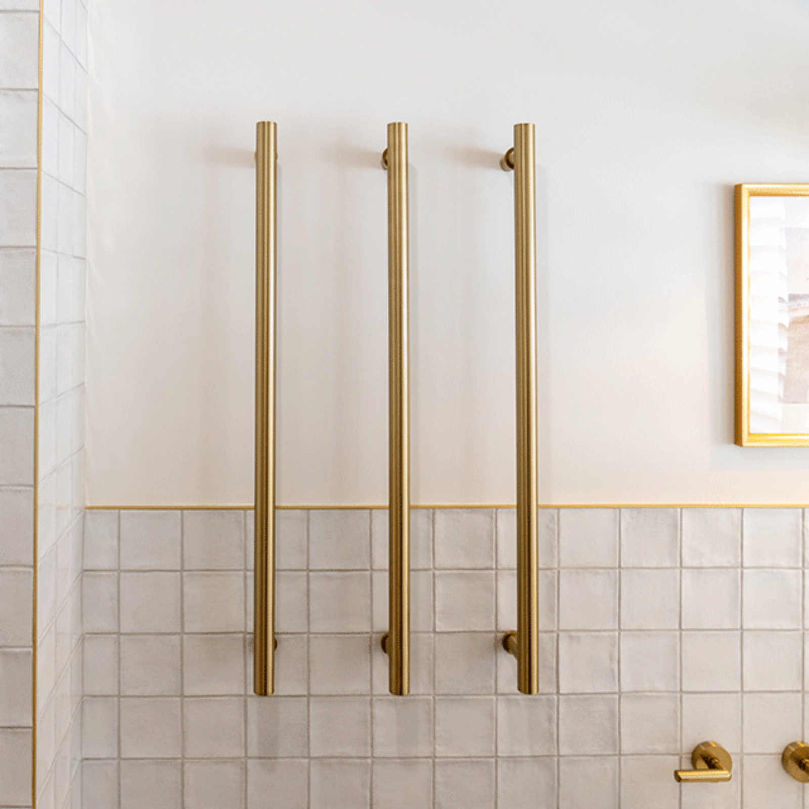 Phoenix heated towel rail in brushed gold, shown vertically mounted on a white tiled bathroom wall with round bars.