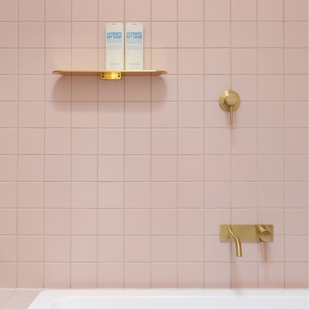 Phoenix Vivid Slimline wall basin/bath mixer set in brushed gold, shown installed on pink tiled wall with shelf and bath edge