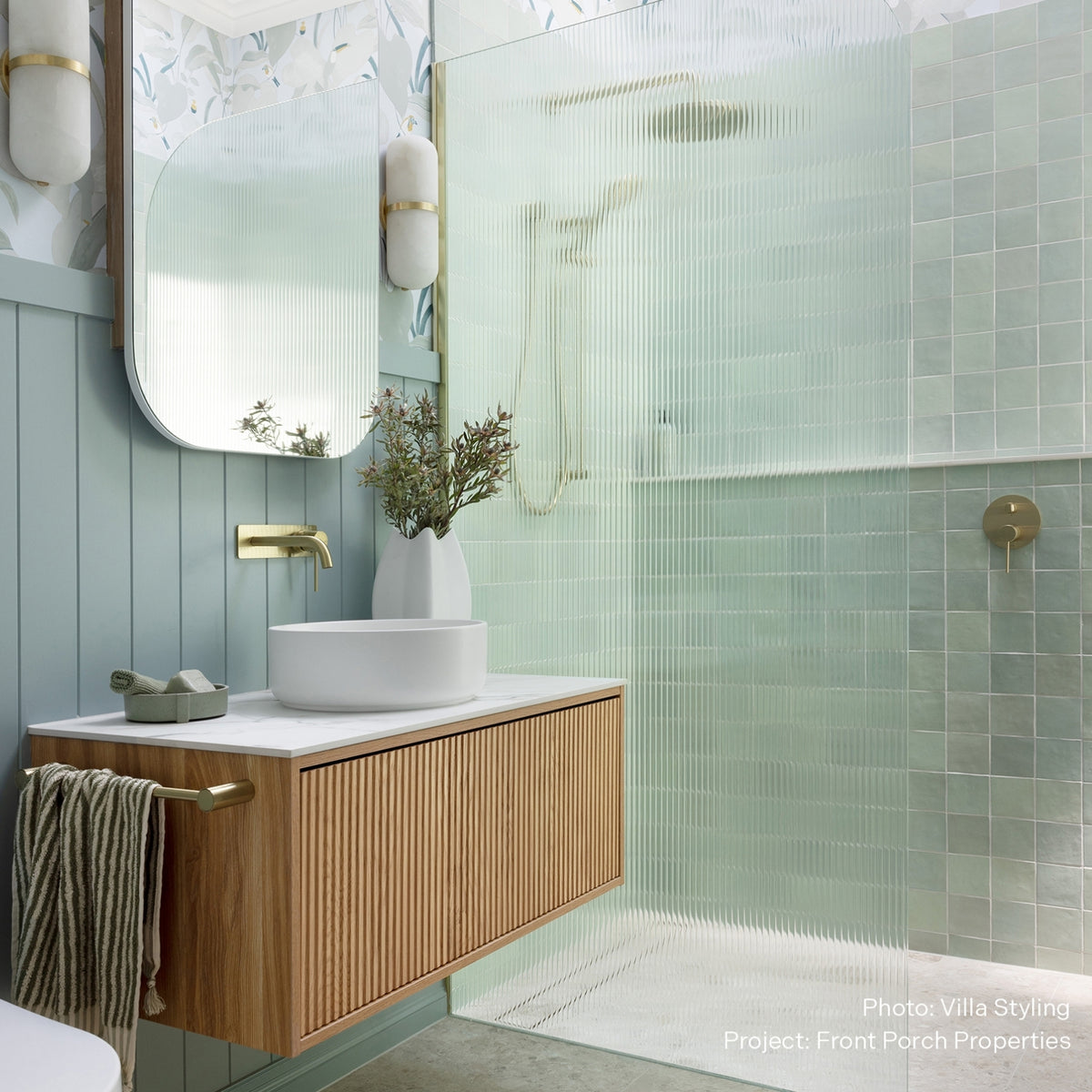 Brushed gold wall basin mixer above a white vanity in a soft green bathroom with fluted glass shower screens