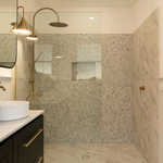 Brushed brass wall-mounted bath/shower mixer in a luxe bathroom, shown beside a shower screen and marble-look tiles.