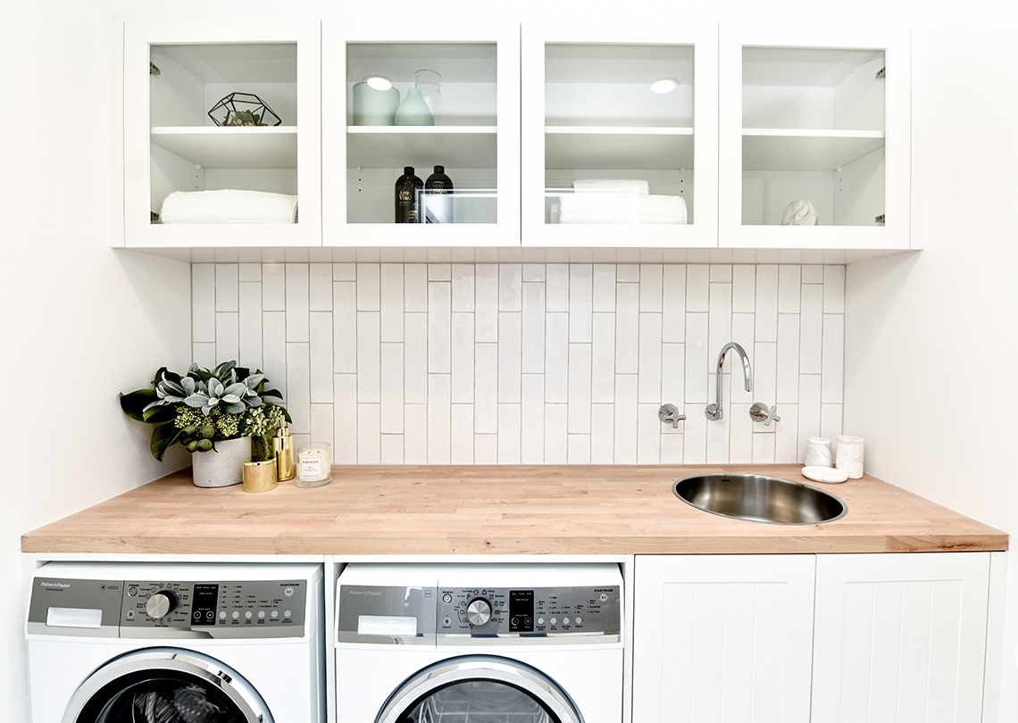 laundry with overhead cupboards, washer and dryer under a wooden counter top and white cupboard complete with round stainless steel sink and chrome wall mounted tap